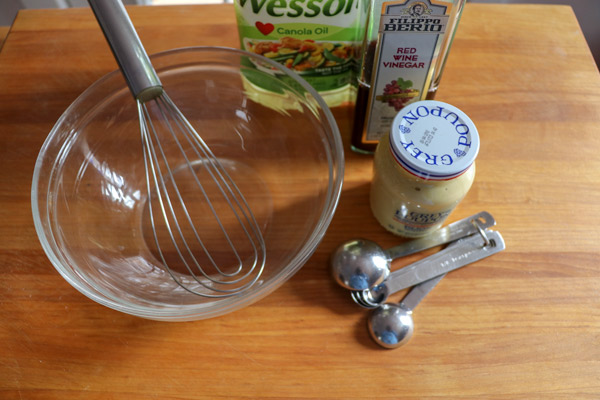 Empty glass bowl and whisk sitting on a wooden cutting board. Canola oil, red wine vinegar, Grey Poupon mustard and measuring sppons are next to the bowl.