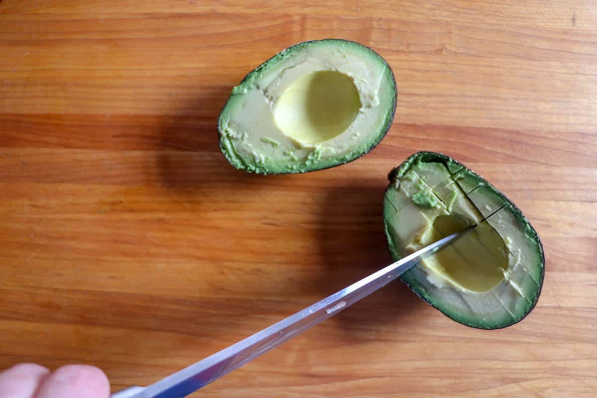 An avocado cut in half on a wooden surface, with a hand using a knife to score the flesh of one half in a grid patternโperfect prep for your Everyday Salad.