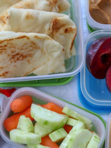 School lunch in containers including naan, cucumbers and carrots, a plum cut in half peanut butter and a bag of Cheetos.