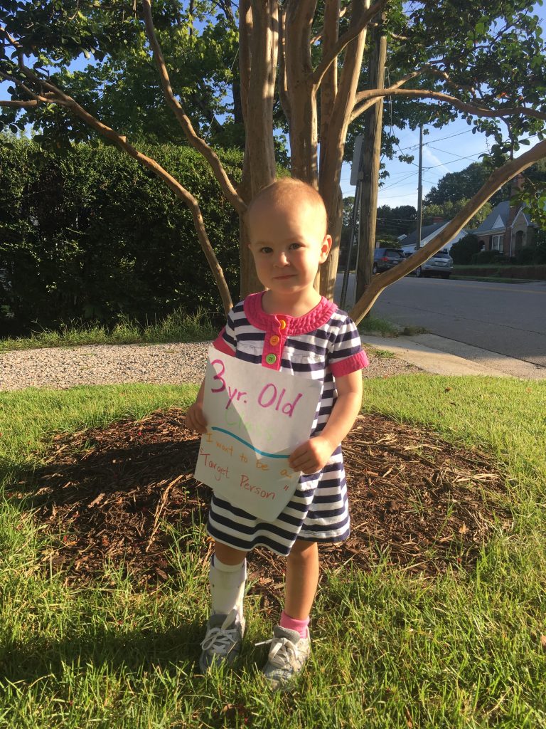 A young girls in a blue and white striped dress holding a first day of school sign for preschool. 