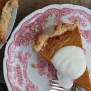 A slice of Thanksgiving pumpkin pie topped with whipped cream on a decorative red and white plate. A fork rests beside the pie. Text above reads, "THE BEST PUMPKIN PIE.
