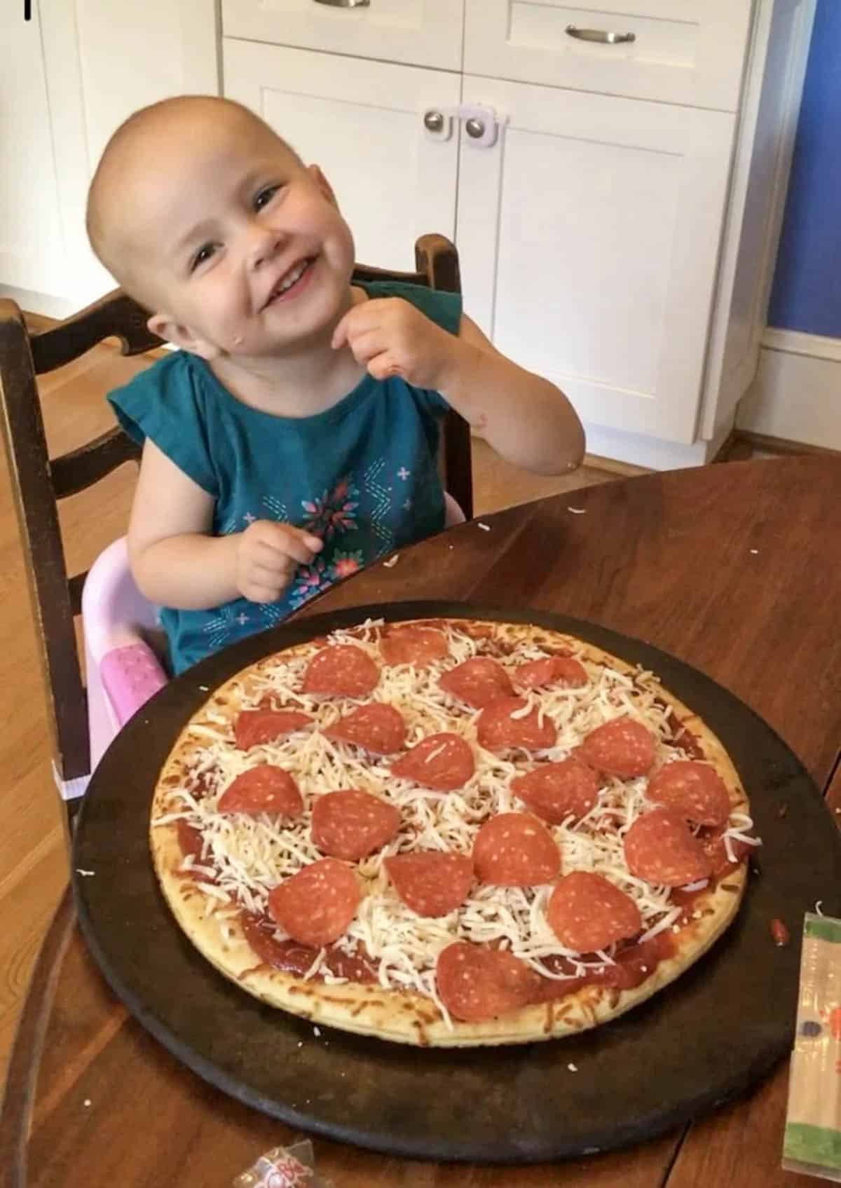 A smiling toddler in a teal dress sits at a table with an uncooked pepperoni pizza—one of her go to meals—on a round tray in front of her, appearing happy and excited in a kitchen setting.