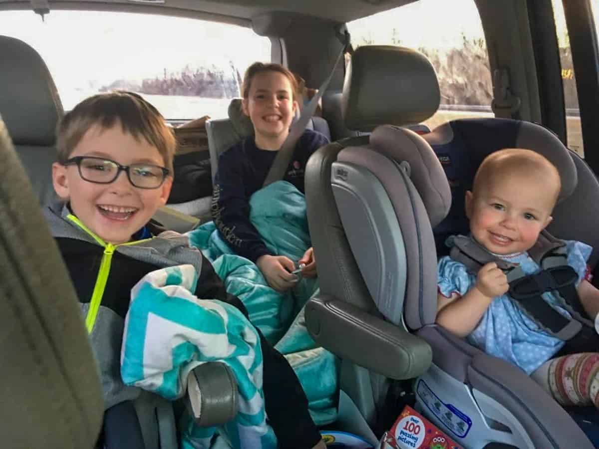 Three children sit in the backseat of a car. Two older kids, wrapped in blankets, smile at the camera. A baby in a car seat beams, holding a toy and wearing blue—ready for adventure or sharing their favorite go to meals on the road.