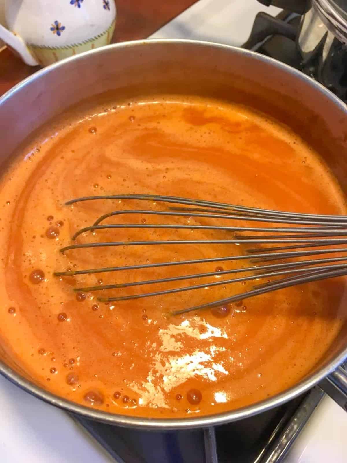 A metal whisk stirs a pot of creamy orange soup on a stovetop, perfect for your go to meals.