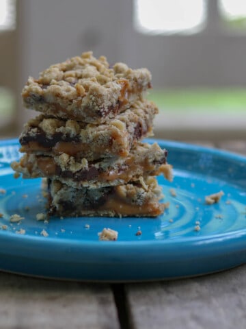 A stack of 4 caramel chocolate oat bars on a bight blue plate sitting on a rustic table.