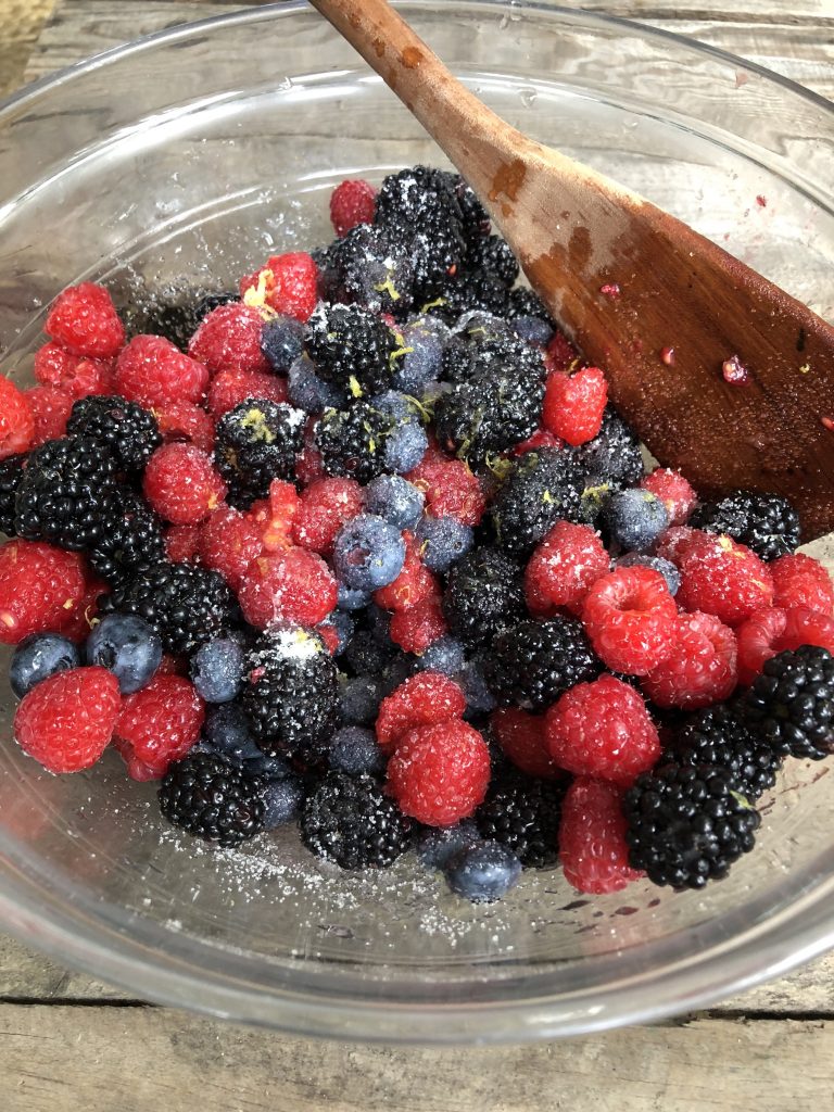 Sugar, lemon zest, lemon juice, and mixed berries in a glass bowl. 