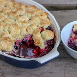 Mixed berry cobbler in a blue and white casserole dish. A portion has been removed and places in a white bowl beside the casserole dish.