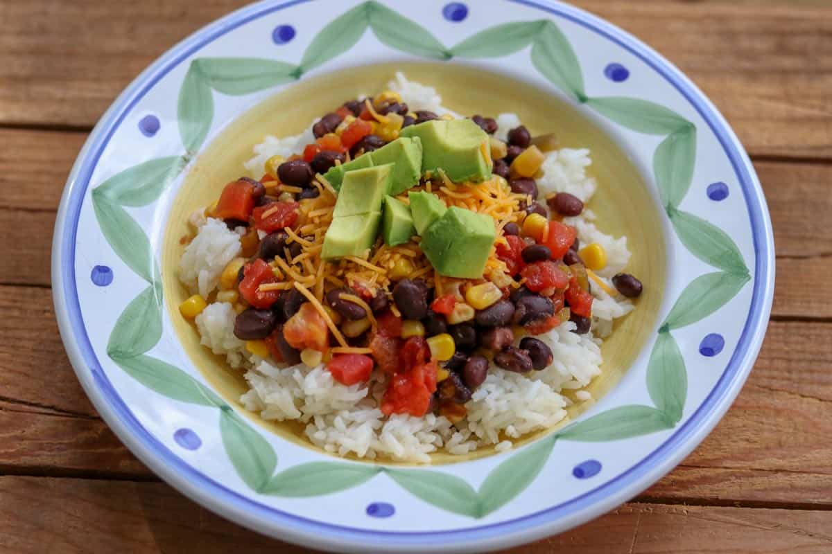 Black beans, corn, tomatoes over rice topped with avocado and cheese and served in a blue and green bowl. 
