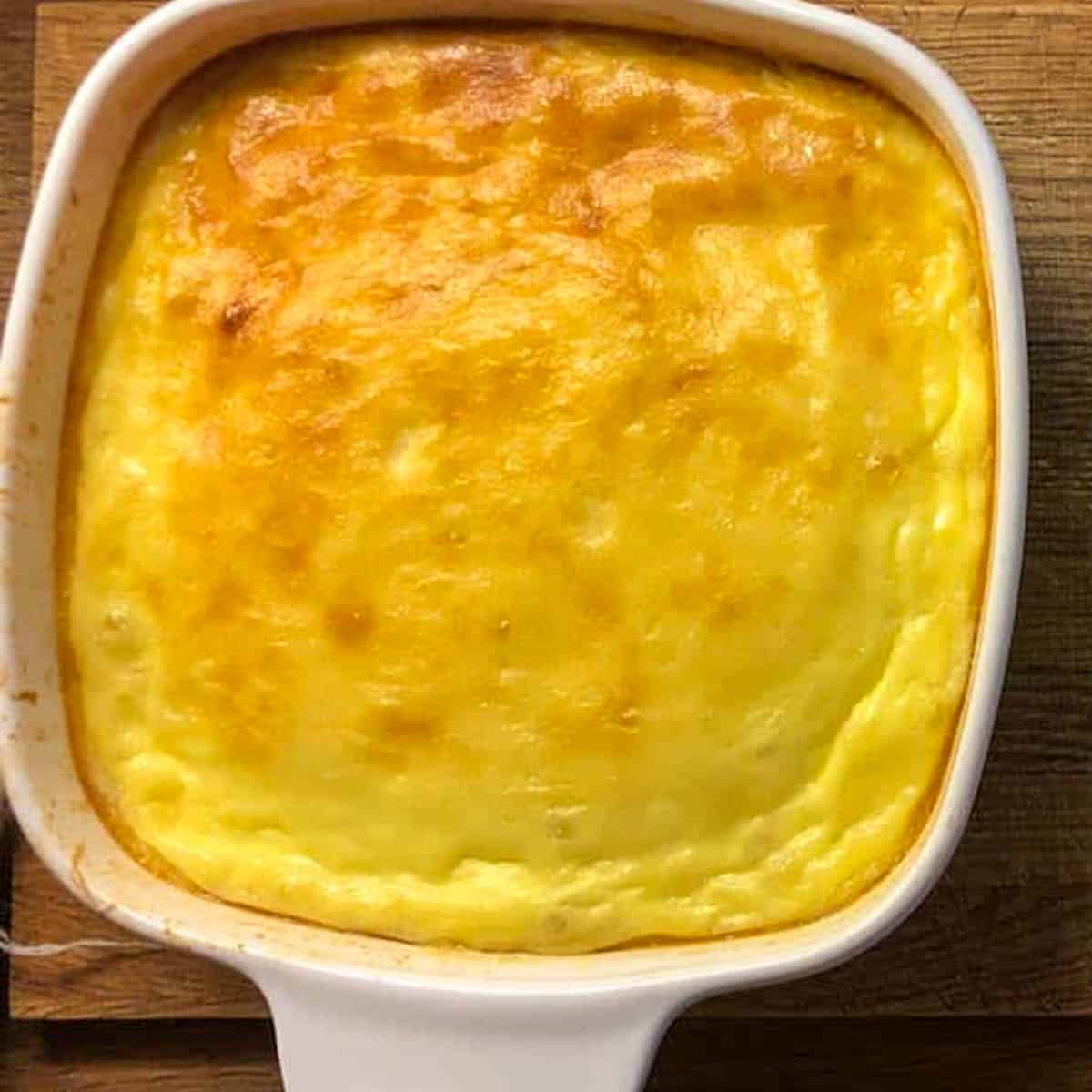 A golden corn pudding sits in a white ceramic baking dish on a wooden surface. Reminiscent of a Kentucky corn pudding, the puddings surface is evenly cooked and slightly puffed, showcasing a smooth, appetizing texture.