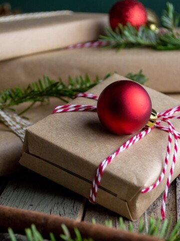 Gifts wrapped in brown wrapping paper, tied with red and white twine and decorated with red Christmas ball and greenery.