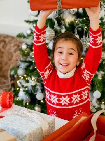 Young girl with brown hair in a red and white snowflake sweater holding a wrapped gift over her head. She's sitting in front of a Christmas tree.