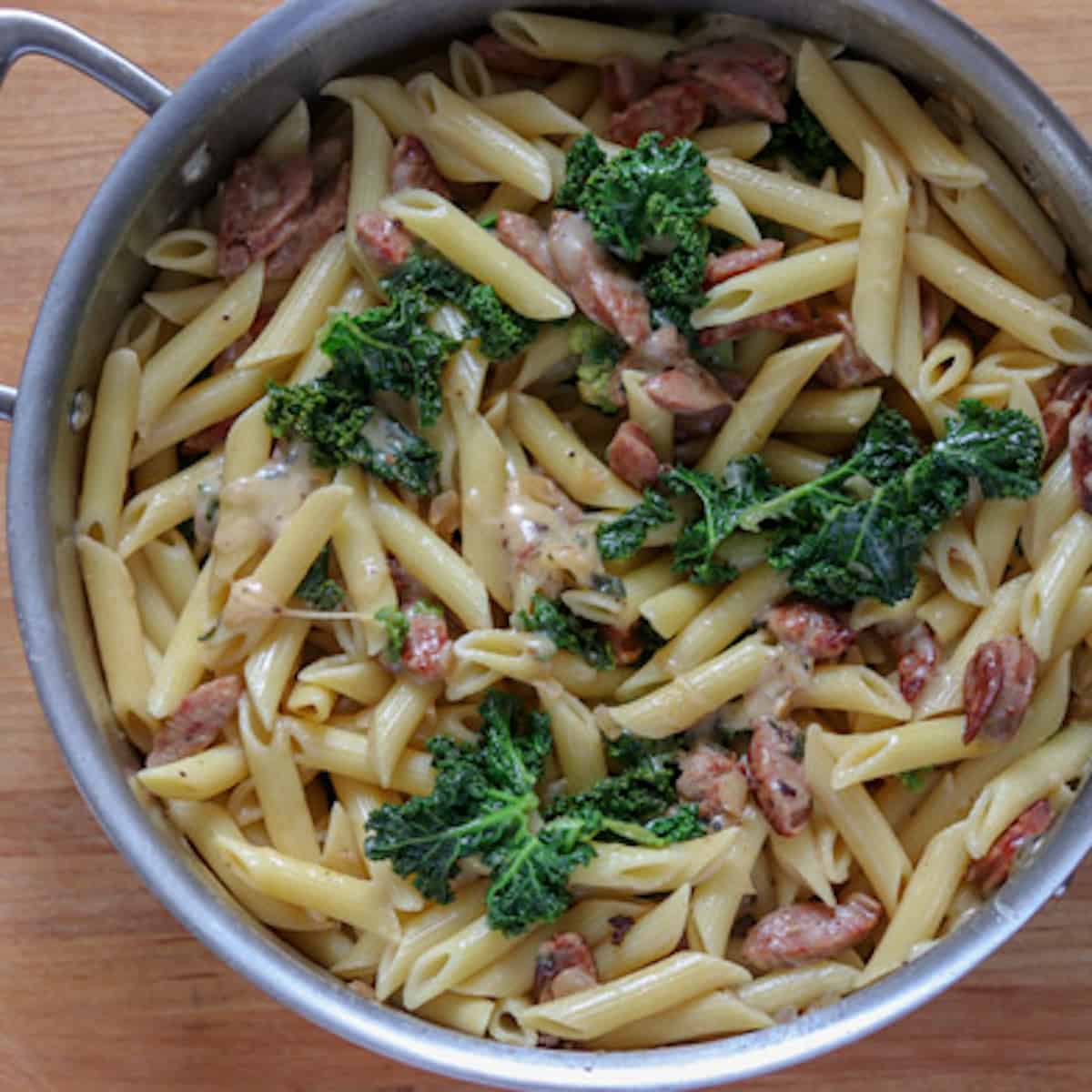 A pot filled with cooked penne pasta mixed with a sage, slices of sausage, and kale, sitting on a wooden surface.