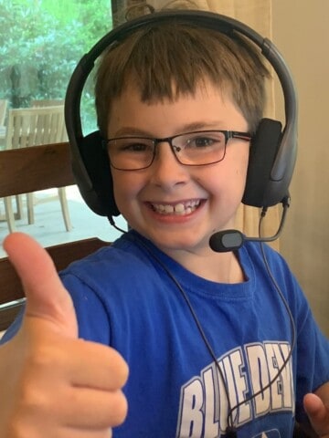 10 year old boy with a blue shirt on gives the thumbs up to the camera. He has on a headset to do virtual school.