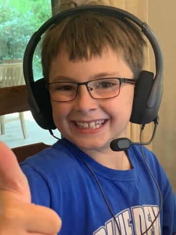 Boy in a blue shirt, glasses, and headphones gives the thumbs up signal.
