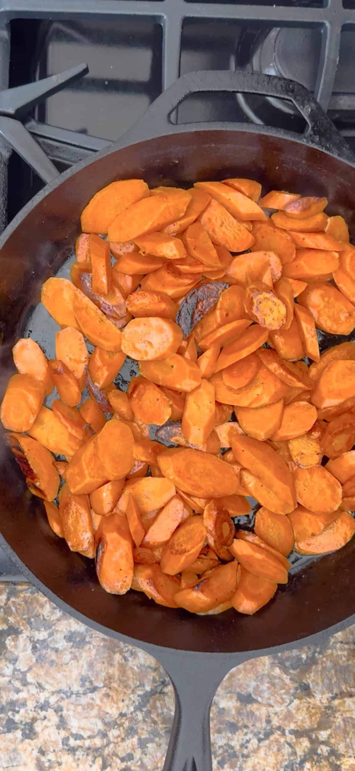 Braised carrots sauté in a black cast iron skillet on a stovetop, with some pieces slightly browned, all resting on a granite countertop.