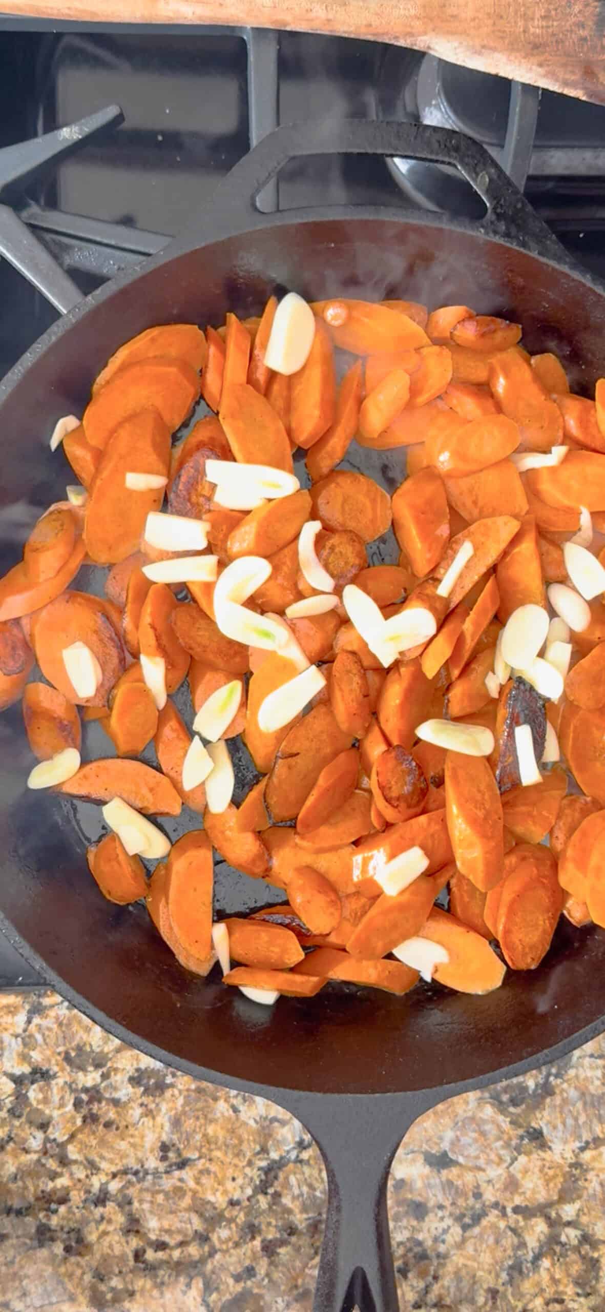 Sliced carrots and garlic cloves are being sautéed in a black cast iron skillet on a stovetop, the perfect start for delicious braised carrots. The vegetables are spread out evenly and just beginning to cook.