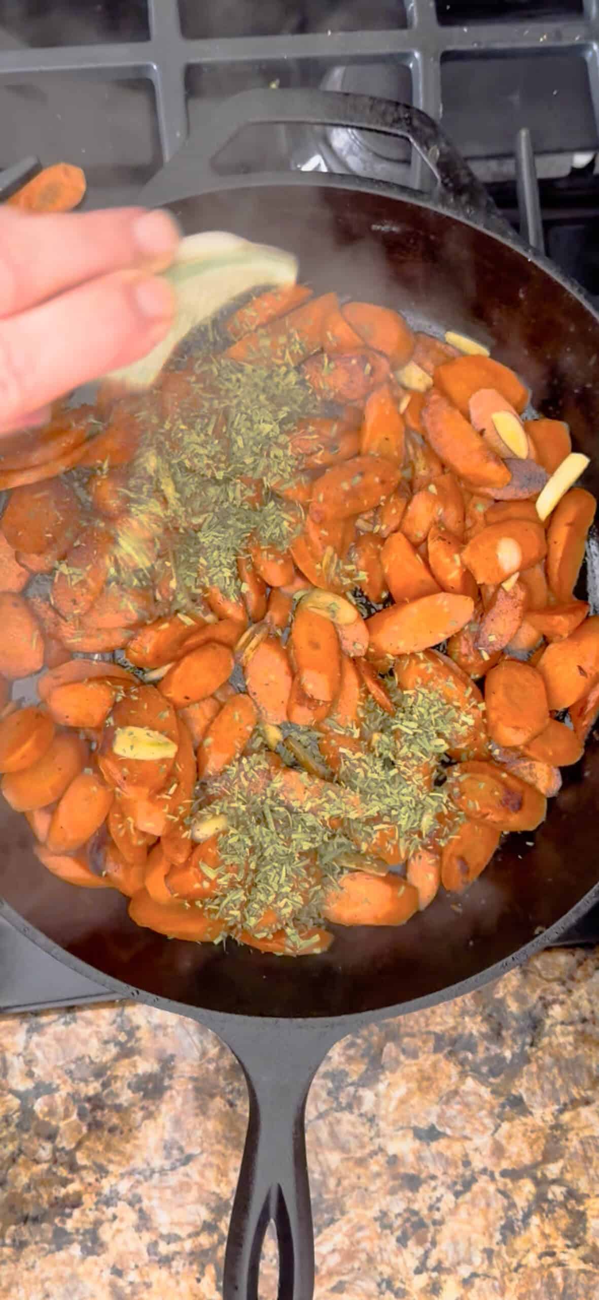 A hand sprinkles herbs over sliced braised carrots and garlic cooking in a black skillet on a stovetop. Steam rises from the mixture, and the skillet sits on a granite countertop.