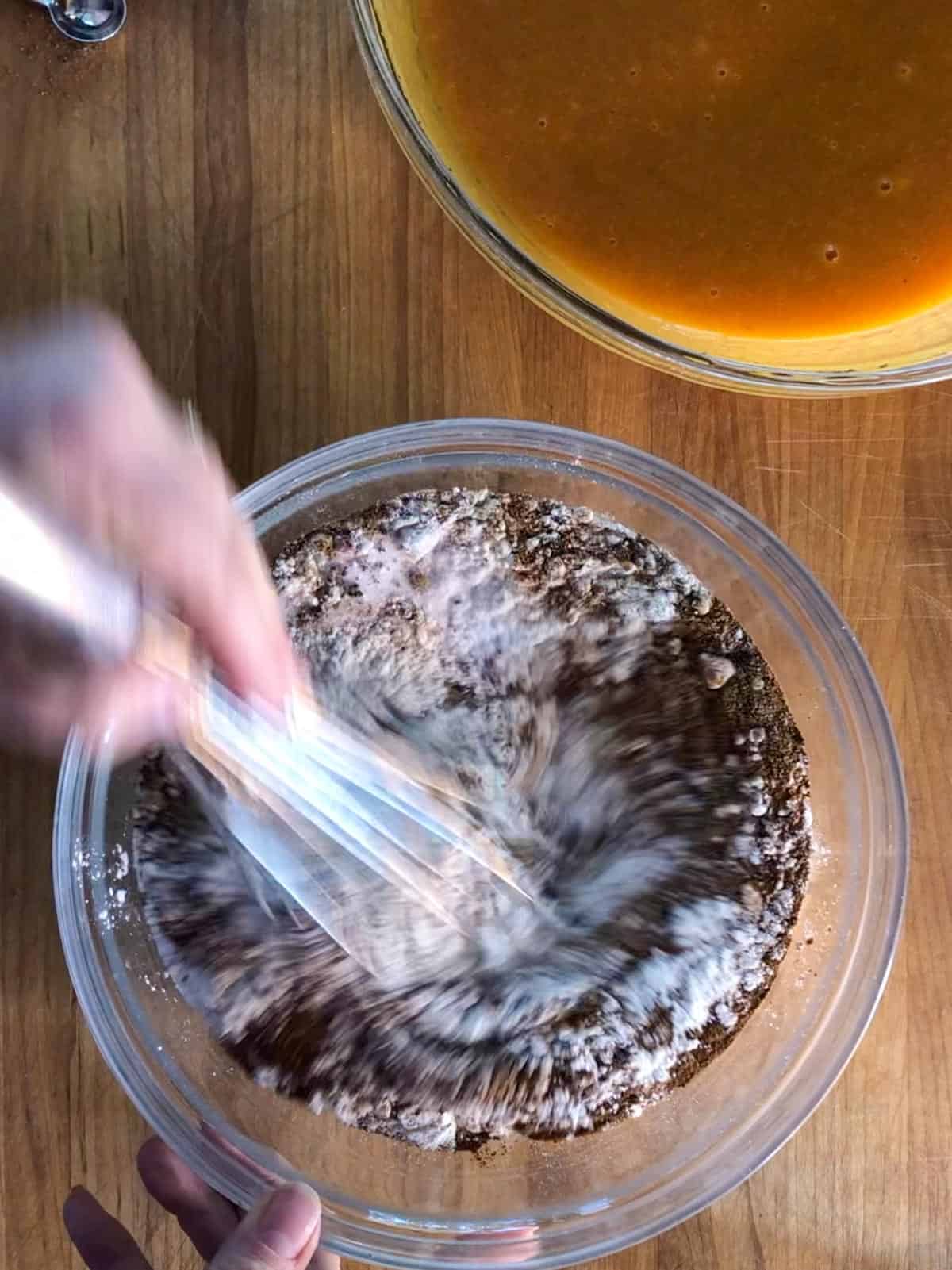 A glass bowl containing the dry ingredients for spiced pumpkin bread. The ingredients are being whisked with a metal whisk. There is also a glass bowl containing the wet ingredients for the spiced pumpkin bread.