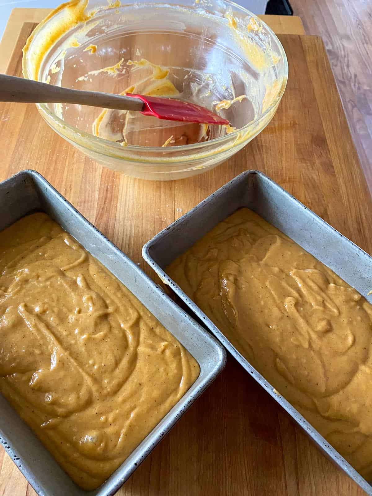 An empty glass bowl and red rubber spatula used to mix spiced pumpkin bread ingredients. And 2 metal bread pans containing pumpkin spice bread batter.