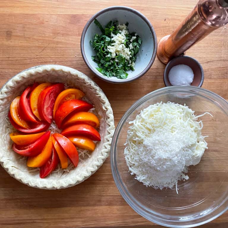 Tomato wedges in an uncooked pie crust next to a bowl of cheeses and a copper pepper grinder.