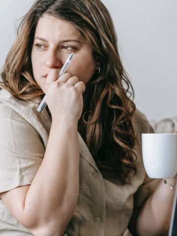 Woman with her hand up to her mouth looking worried. Coffee cup is in her other hand.
