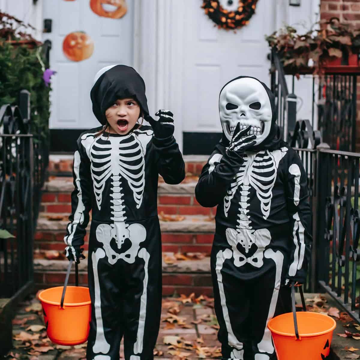 2 children in skeleton costumes, one with a skeleton mask on their face. Each child is holding an orange halloween candy bucket.