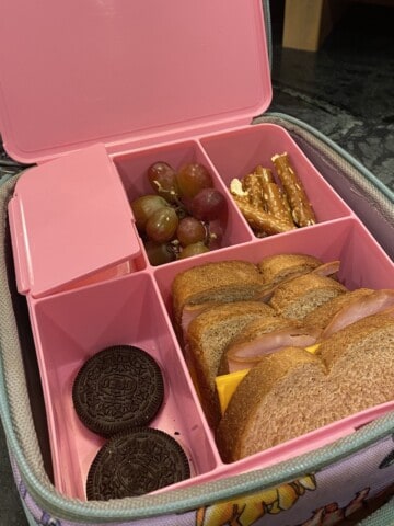 Pink bentobox lunchbox filled with a sandwich, pretzels, grapes and 2 oreos.