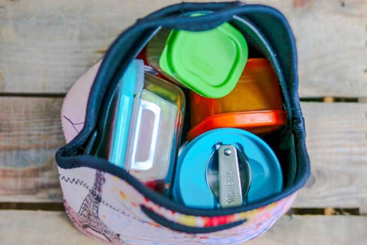 A top-down view of lunch box essentialsโa lunch bag filled with colorful food containers and a metal utensil, all neatly arranged inside the bag, placed on a wooden surface.