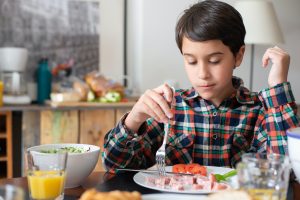 Boy with brown hair and plaid shirt sitting at a table holding is fork in preparation to eat. 