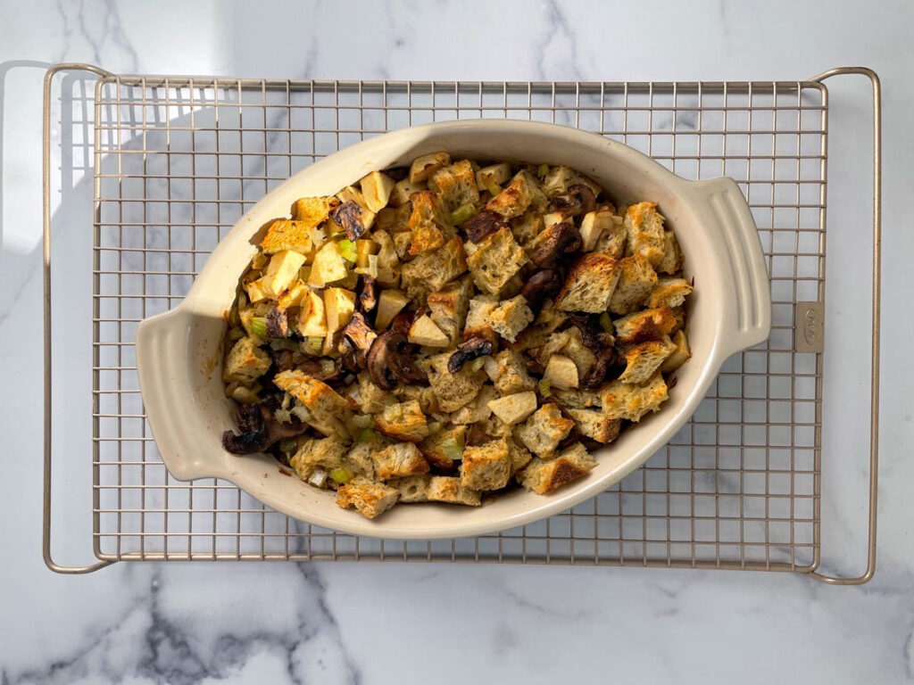 Baked stuffing with toasted bread cubes in an oval dish sitting on a gold colored cooling rack on a marble counter.