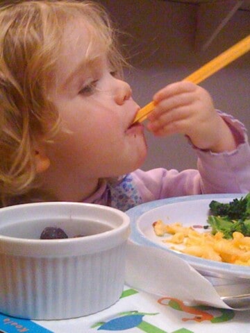 Girl with light wavy hair eats with orange chopsticks. A plate of broccoli andmacaroni and cheese, and bowl of grapes sit in front of her.