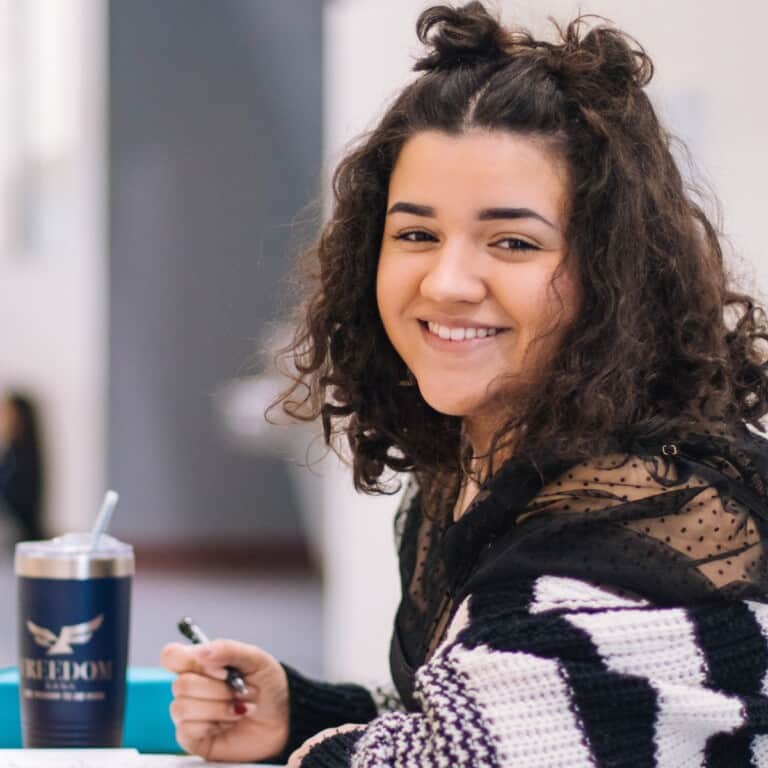 Girl with dark wavy hair sits at a table studying with a water bottle. She smiles at the camera.