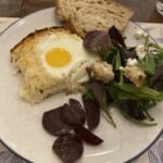 A plate with a baked egg on rosti casserole, mixed green salad with croutons and cheese, sliced beets, and a slice of rustic bread.