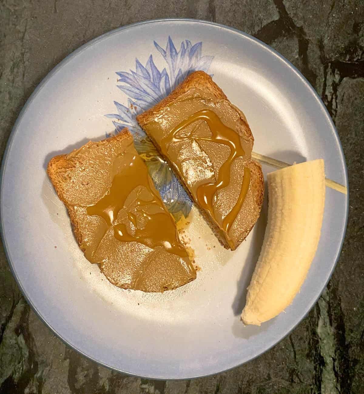 A plate with two slices of toast topped with peanut butter and a half-peeled banana on the side, set on a dark stone surface—perfect for quick after school snacks.