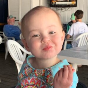 3 year old in light blue bathing suit top eating a piece of fried shrimp, making a face like she's not so sure she likes it.