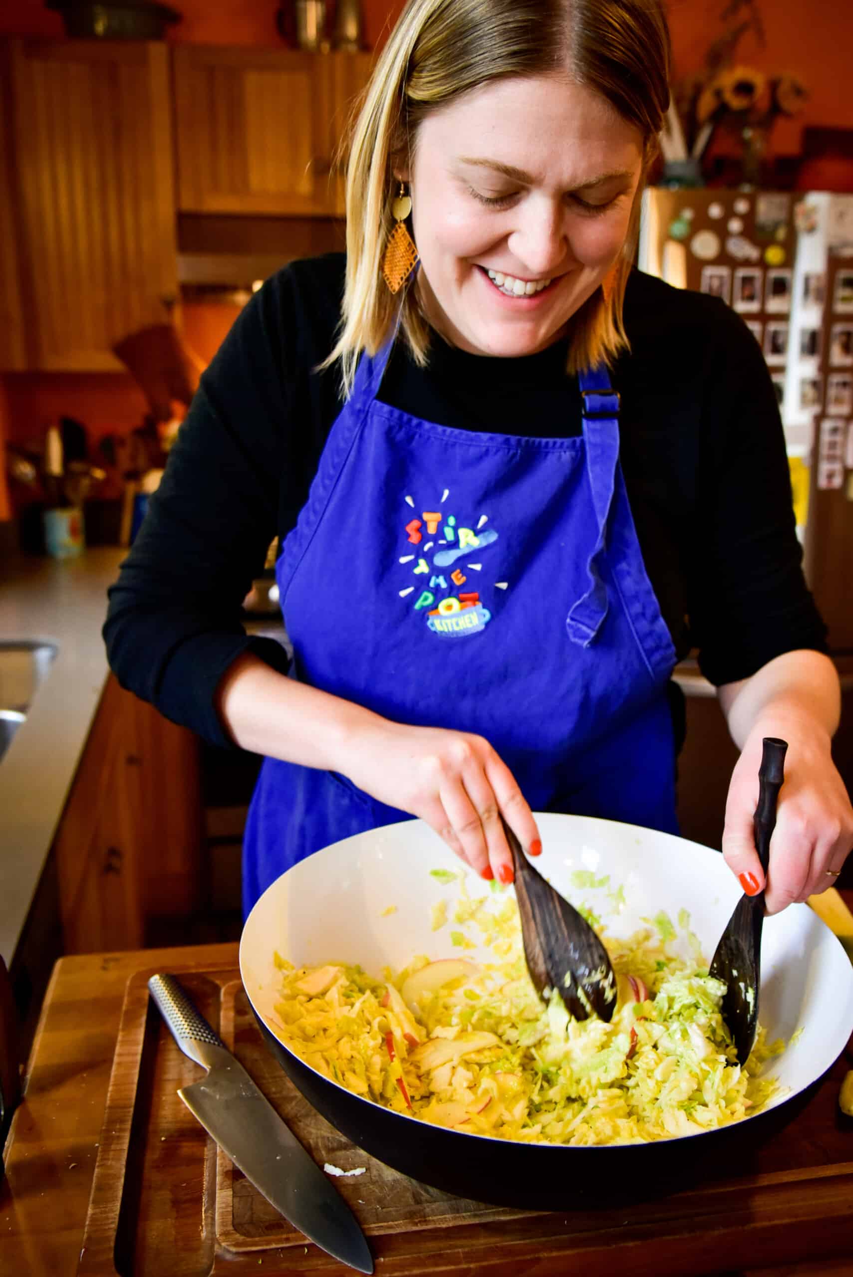 Woman with mid-length blond hair, wearing a blue apron, tosses a salad in a big salad bowl. 