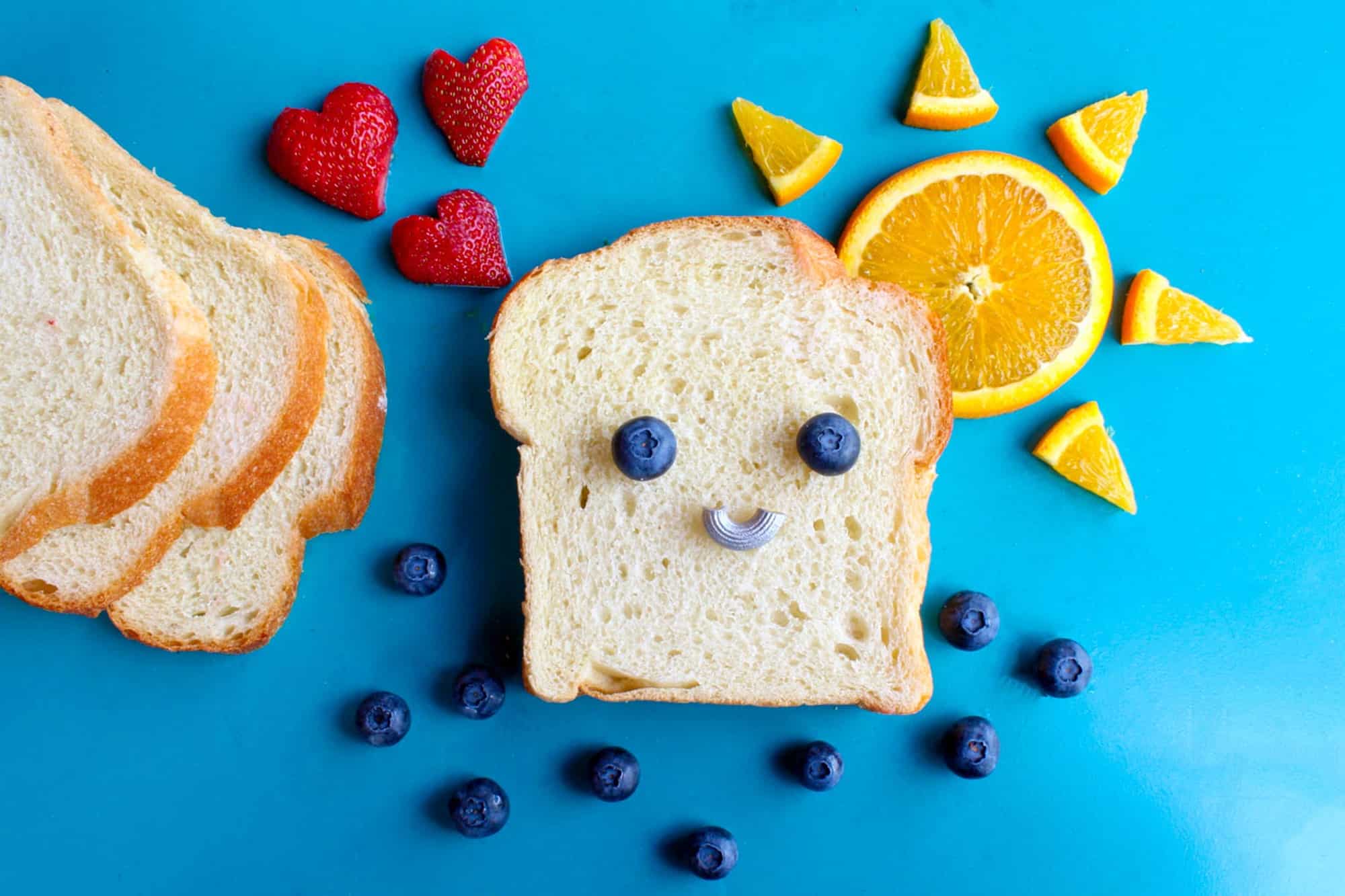 Blue background with a slice of bread with blueberries make a smile on the bread. There is a sunshine to the right of the slice of bread made of orange slices and heart shaped strawberries to the right of the slice. 
