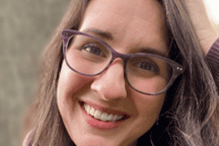 Woman with brown hair and glasses, wearing a lavender sweater, smiles for the camera.