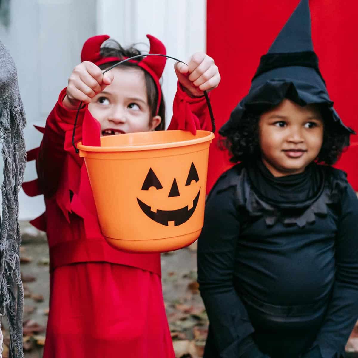 2 young kids, one dressed as a devil and one dressed as a witch. The child dressed as a devil is holding up a Halloween pumpkin candy bucket. 