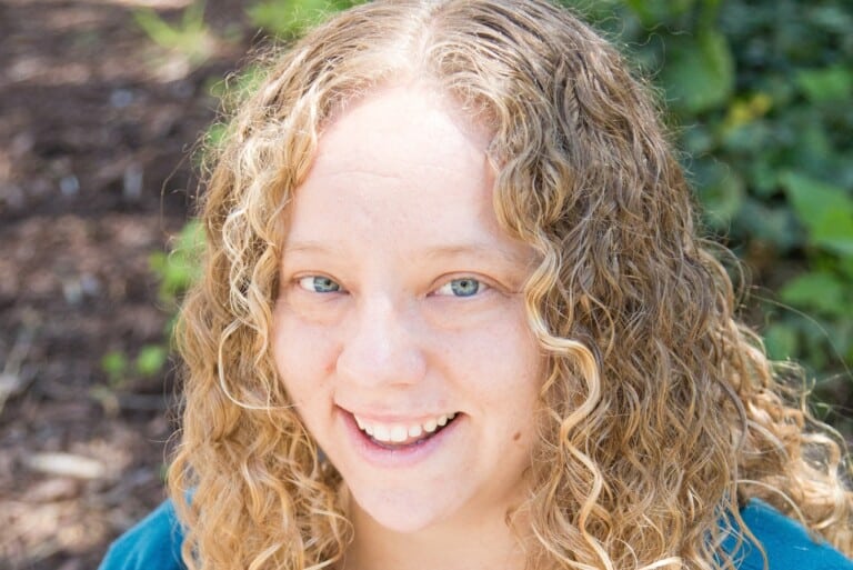 Woman with blond curly hair and a blue cardigan sweater smiles for the camera.