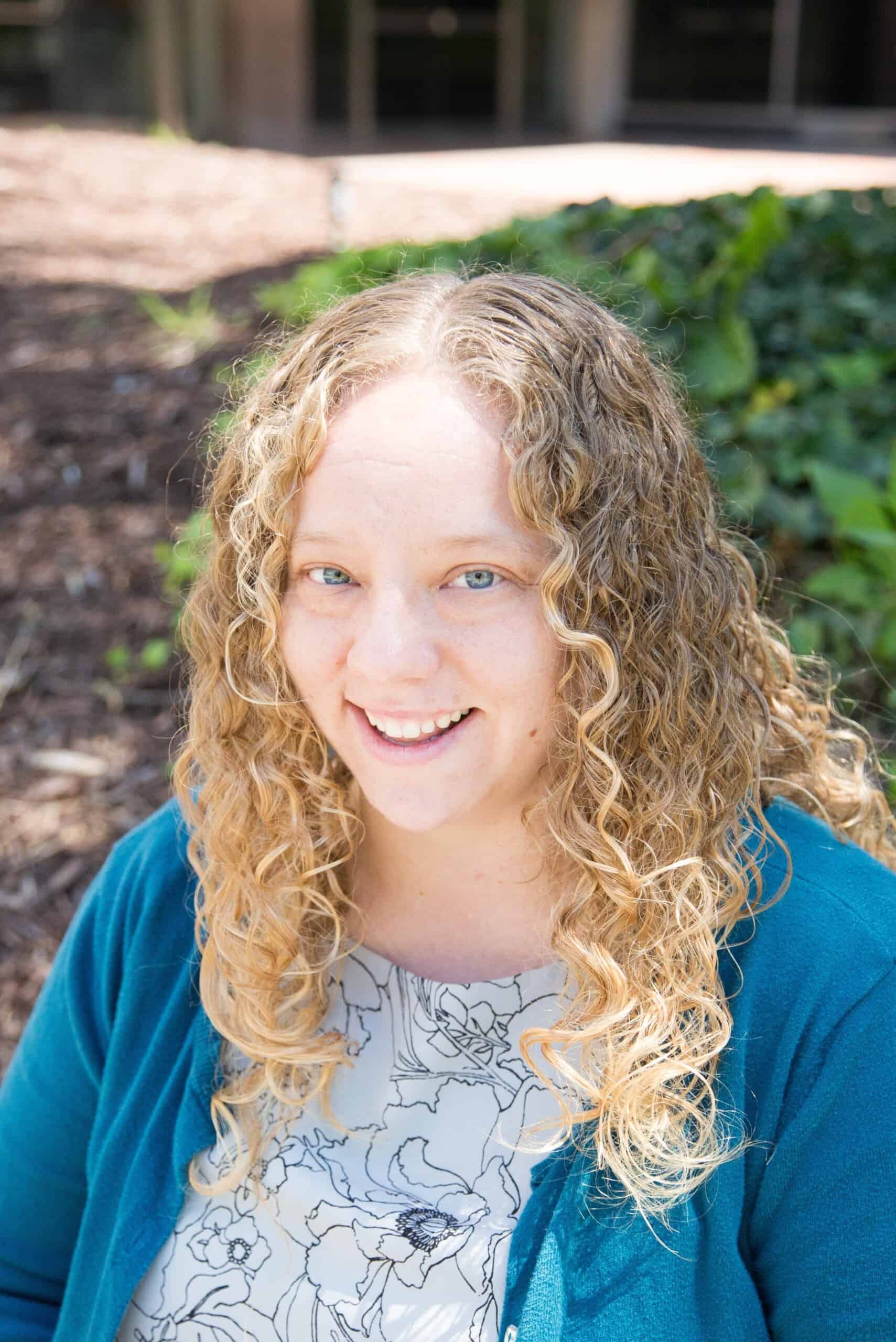 Woman with blond curly hair and a blue cardigan sweater smiles for the camera. 