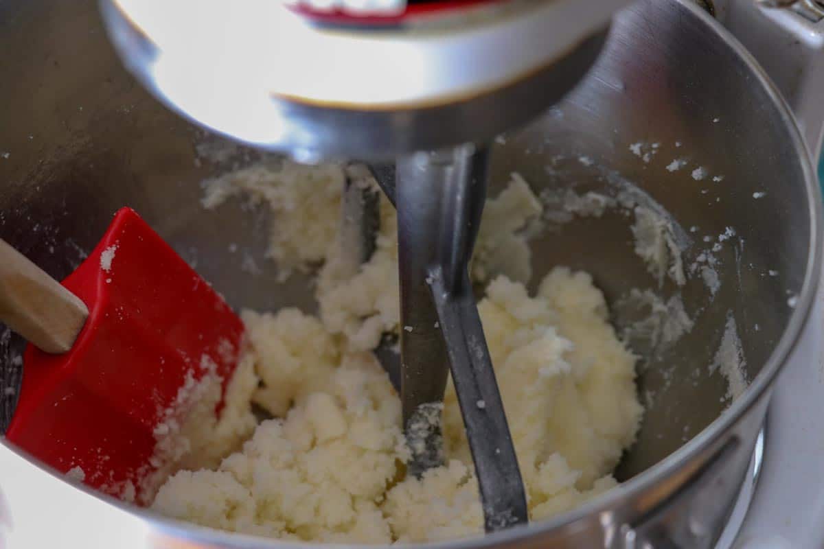 Red rubber spatula scraping down the sides of the butter sugar mixture in a Kitchen Aid stand mixer bowl.