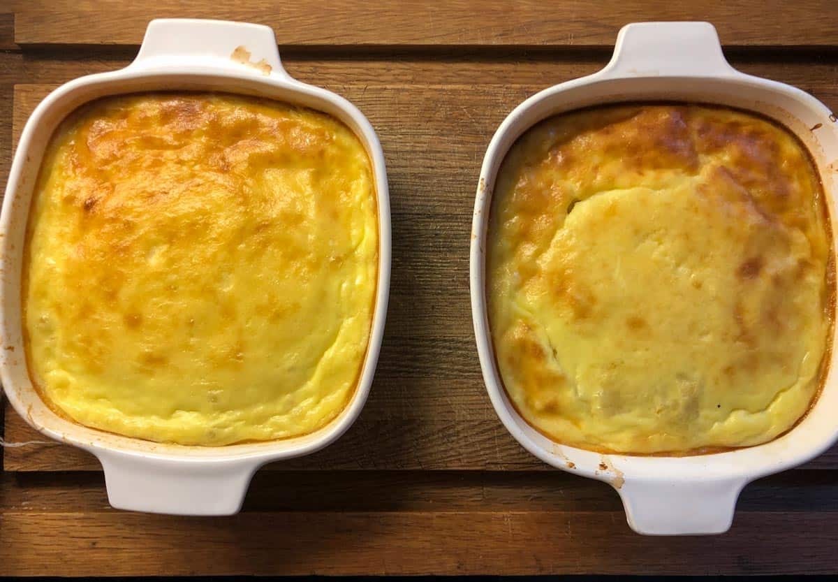 Two square baking dishes of baked corn pudding sitting on a wooden cutting board. 