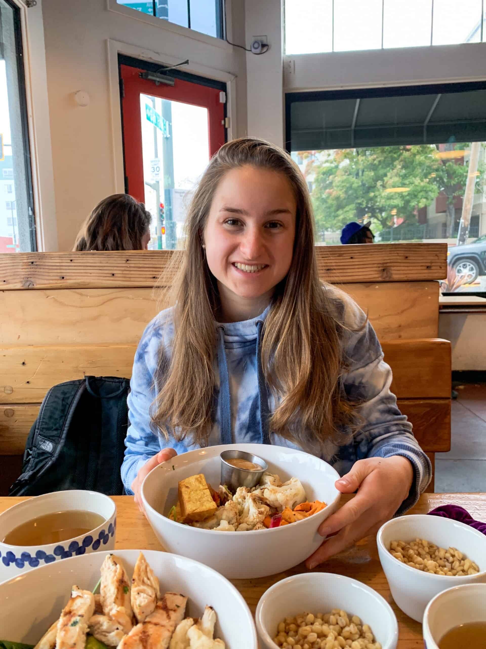Young woman with long blond hair sits at a table with several bowls of food. She smiles for the camera. 