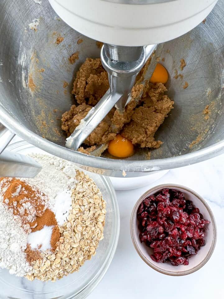 Eggs in a stand mixer bowl with butter and sugar mixture. Dry ingredients and craisins for the oatmeal craisin cookie dough are next to the mixer bowl.