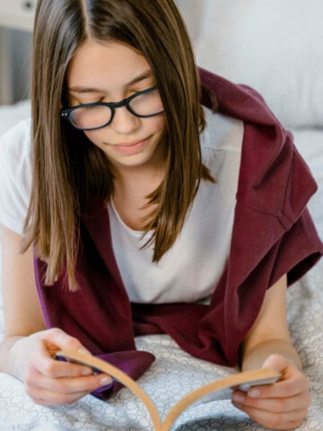 Teenage female with long brown hair and glasses lying on her bed reading a book.