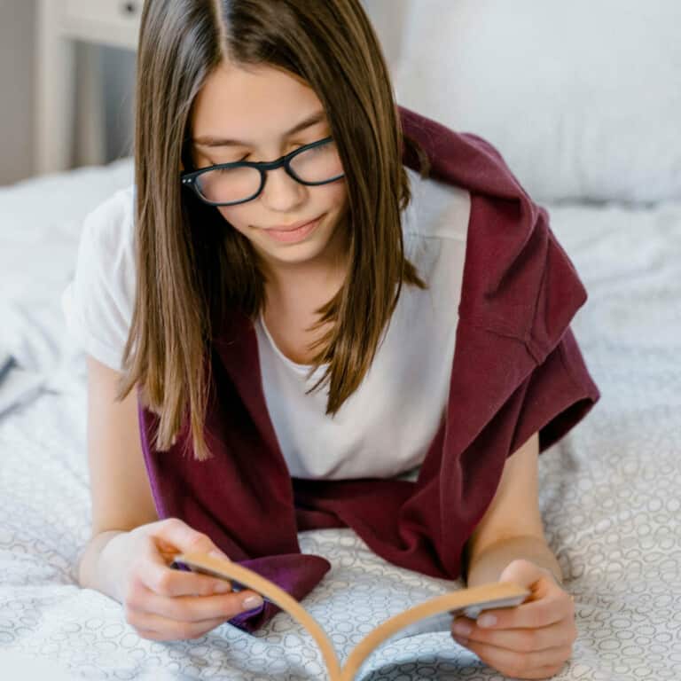 Teenage female with long brown hair and glasses lying on her bed reading a book.