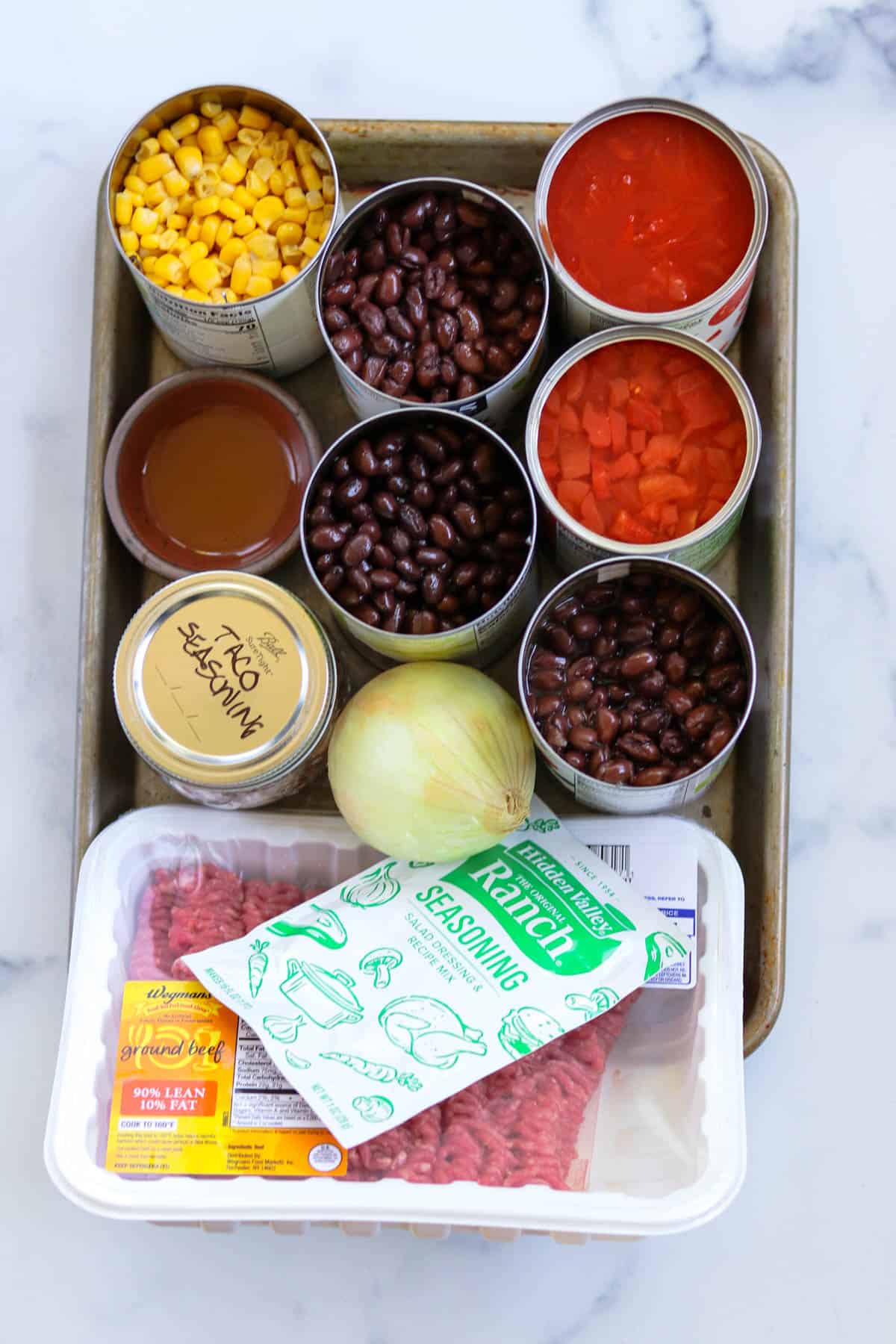 Ingredients to make easy black bean taco soup: canned corn, canned black beans, canned diced tomatoes, olive oil, onion, taco seasoning, ranch dressing packet, and ground beef gathered together on a quarter sheet pan.