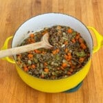 A yellow pot filled with an easy lentil dish, also chopped carrots, onions, and celery. A wooden spoon rests inside the pot, which sits on a wooden surface.