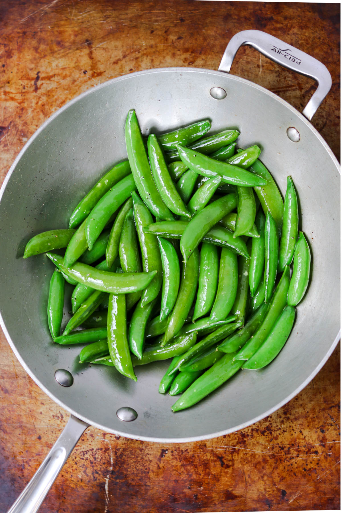 A stainless steel pan filled with a veggie side of green sugar snap peas, lightly sautéed. The peas glisten, reflecting light, and the background is a warm, textured brown surface. The pan handle is visible on the left-hand side.